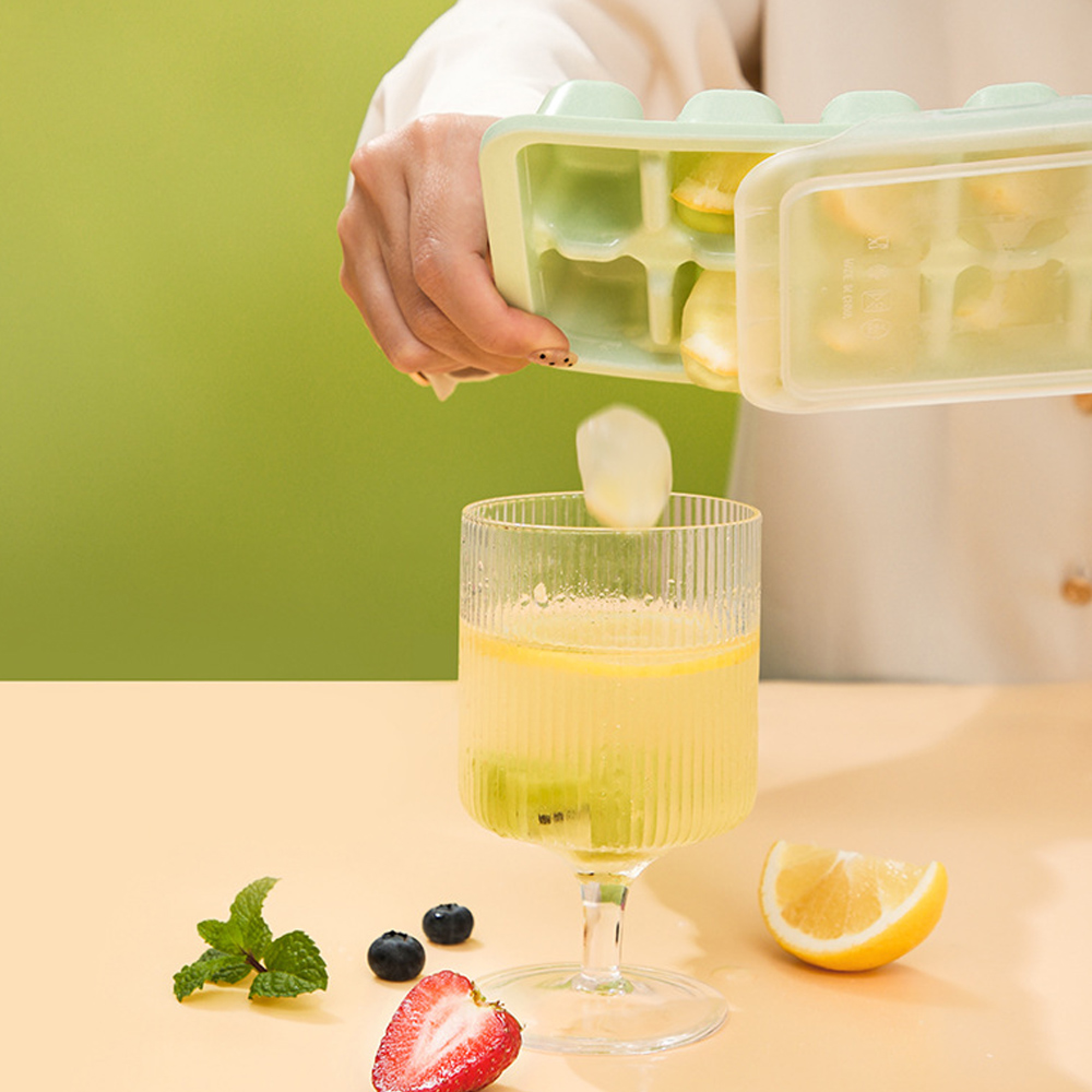 Eine Hand gibt Eiswürfel mit Zitronen in ein Glas mit Getränk. Beeren, Minze und Zitrone liegen daneben.