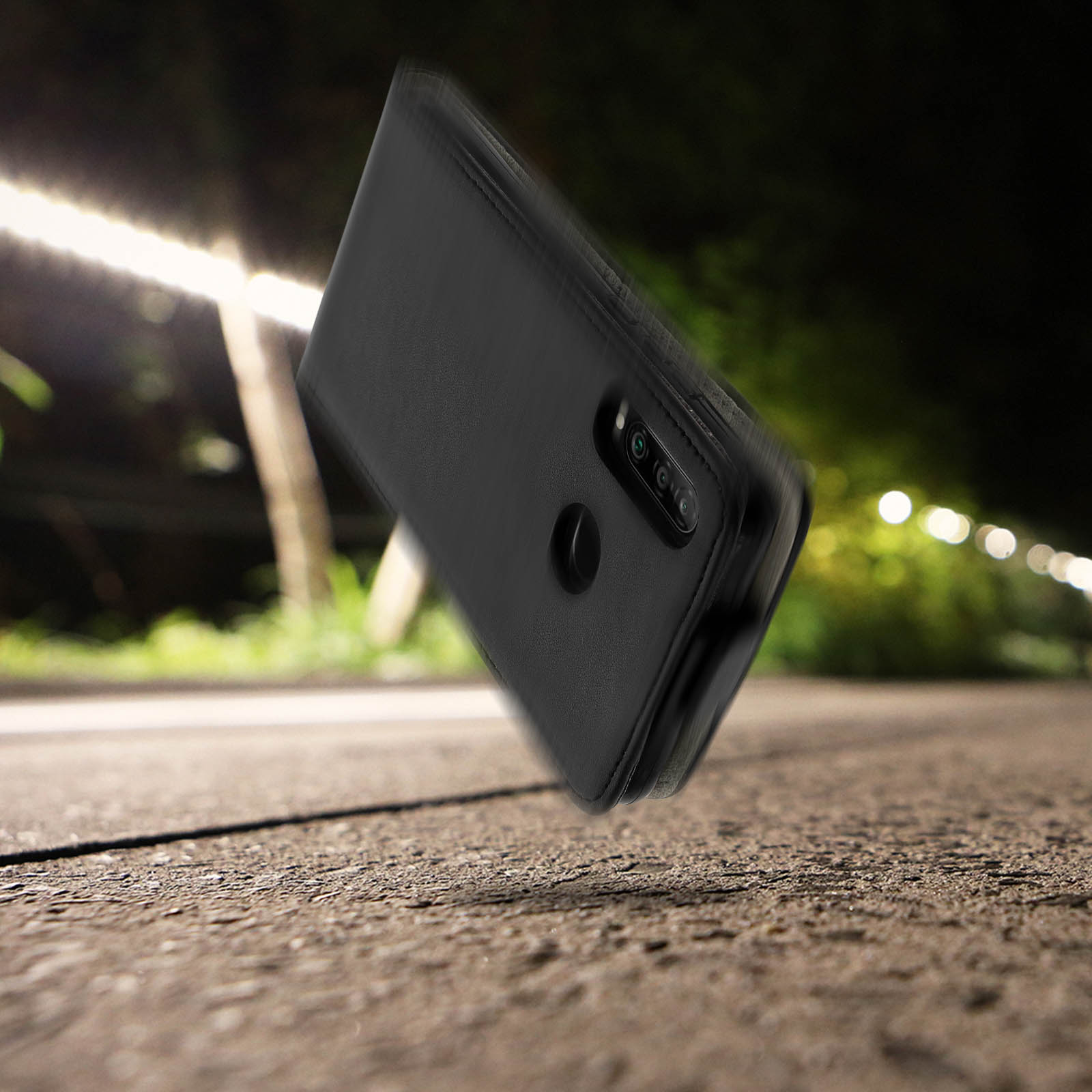 Funda de teléfono negra cayendo sobre asfalto, calle nocturna con luces al fondo.