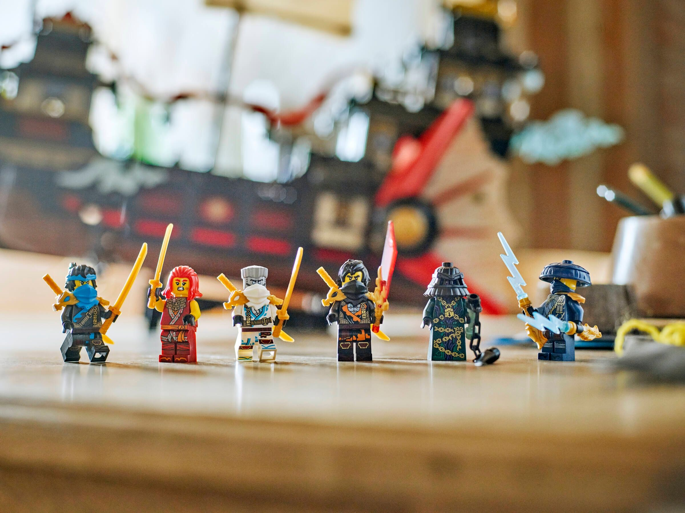 Sechs Lego-Ninja-Figuren mit Schwertern stehen auf einer Holzoberfläche. Ein Lego-Schiff ist im Hintergrund unscharf.