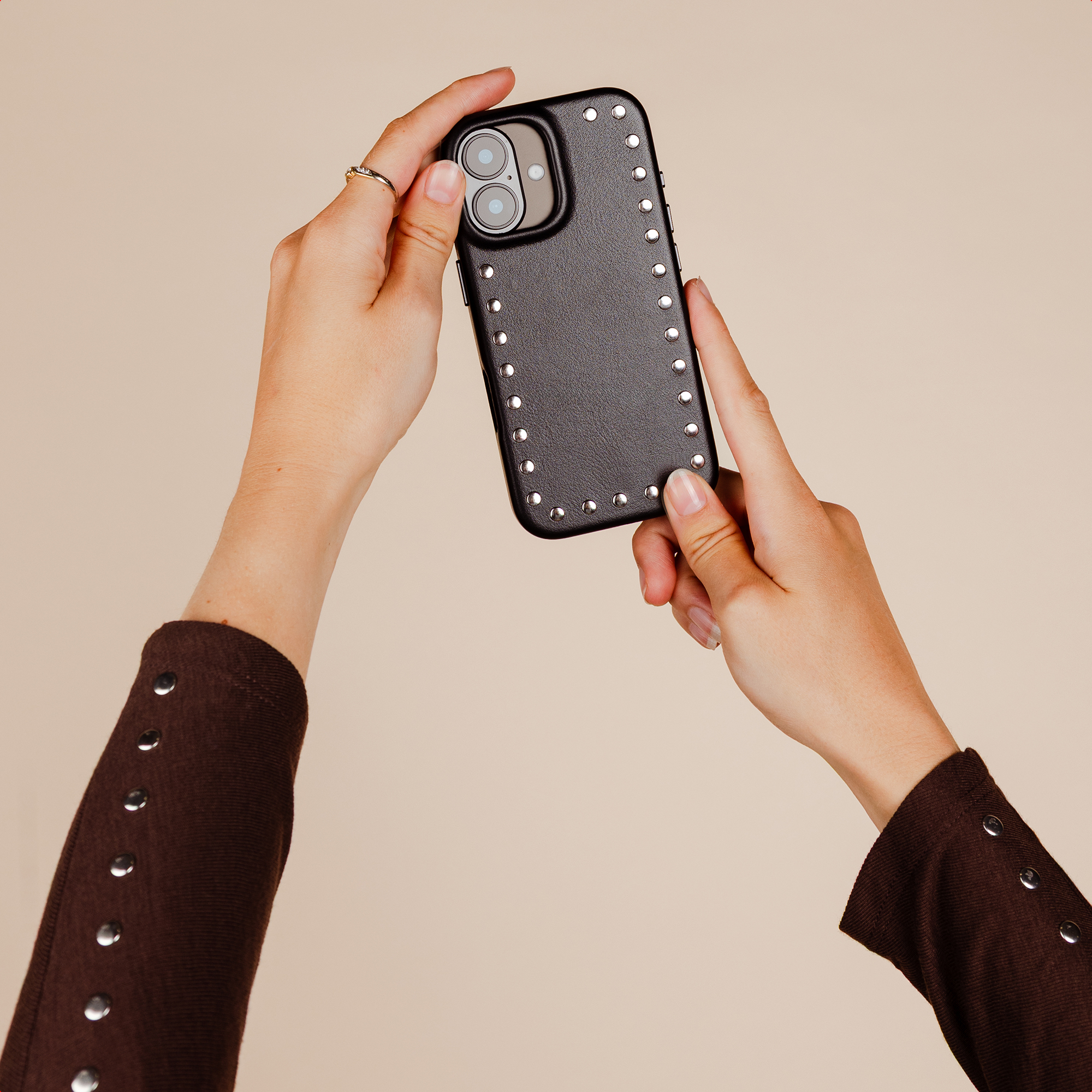Hands hold a black phone with studs and two cameras. Brown shirt.