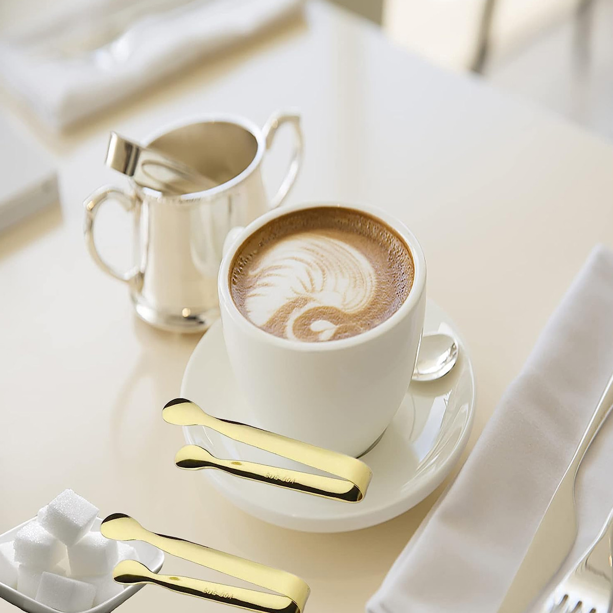 Kaffee mit Latte Art, Zuckerdose, Würfelzucker und Zuckerzange auf einem Tisch.