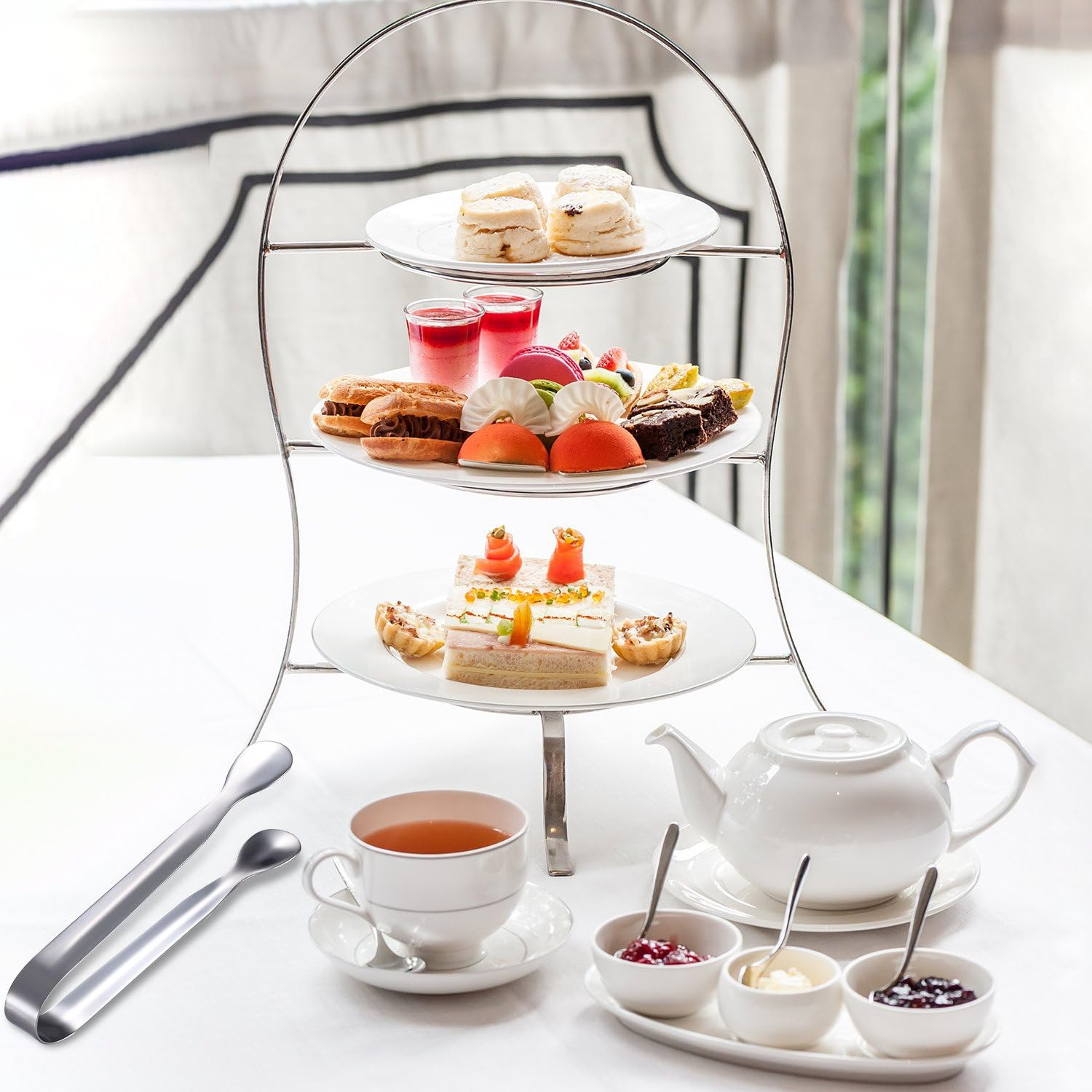 Ein dreistöckiger Ständer mit verschiedenen Gebäckstücken, Sandwiches und Scones. Teekanne, Tasse und Zange auf dem Tisch.