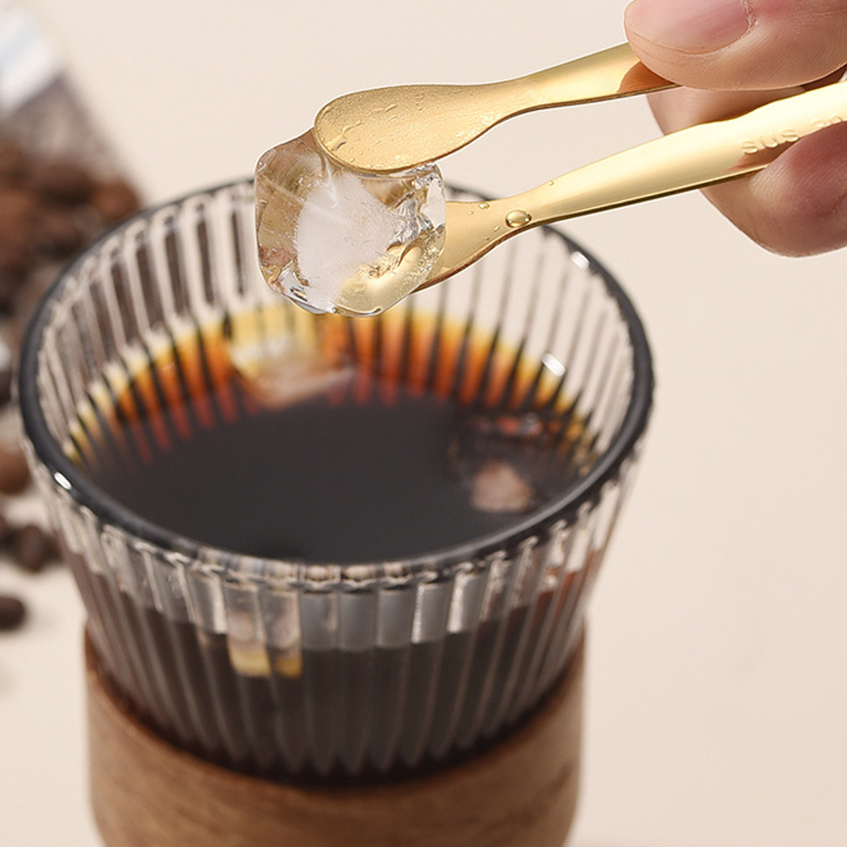 Goldene Zange hält einen Eiswürfel über ein Glas dunklen Kaffees. Kaffeebohnen im Hintergrund.