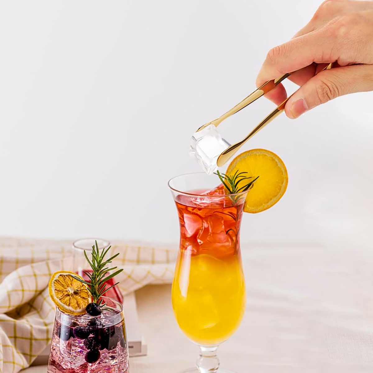 Eine Hand benutzt eine goldene Zange, um ein Eiswürfel in einen geschichteten Cocktail zu geben.