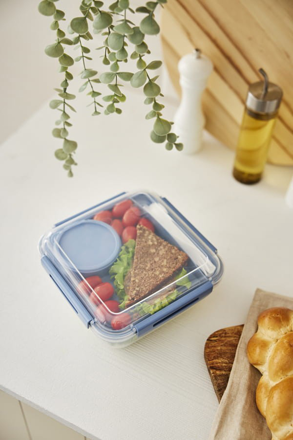 Pudełko na lunch z kanapką, pomidorkami i sałatą, obok chleba i butelki z oliwą.