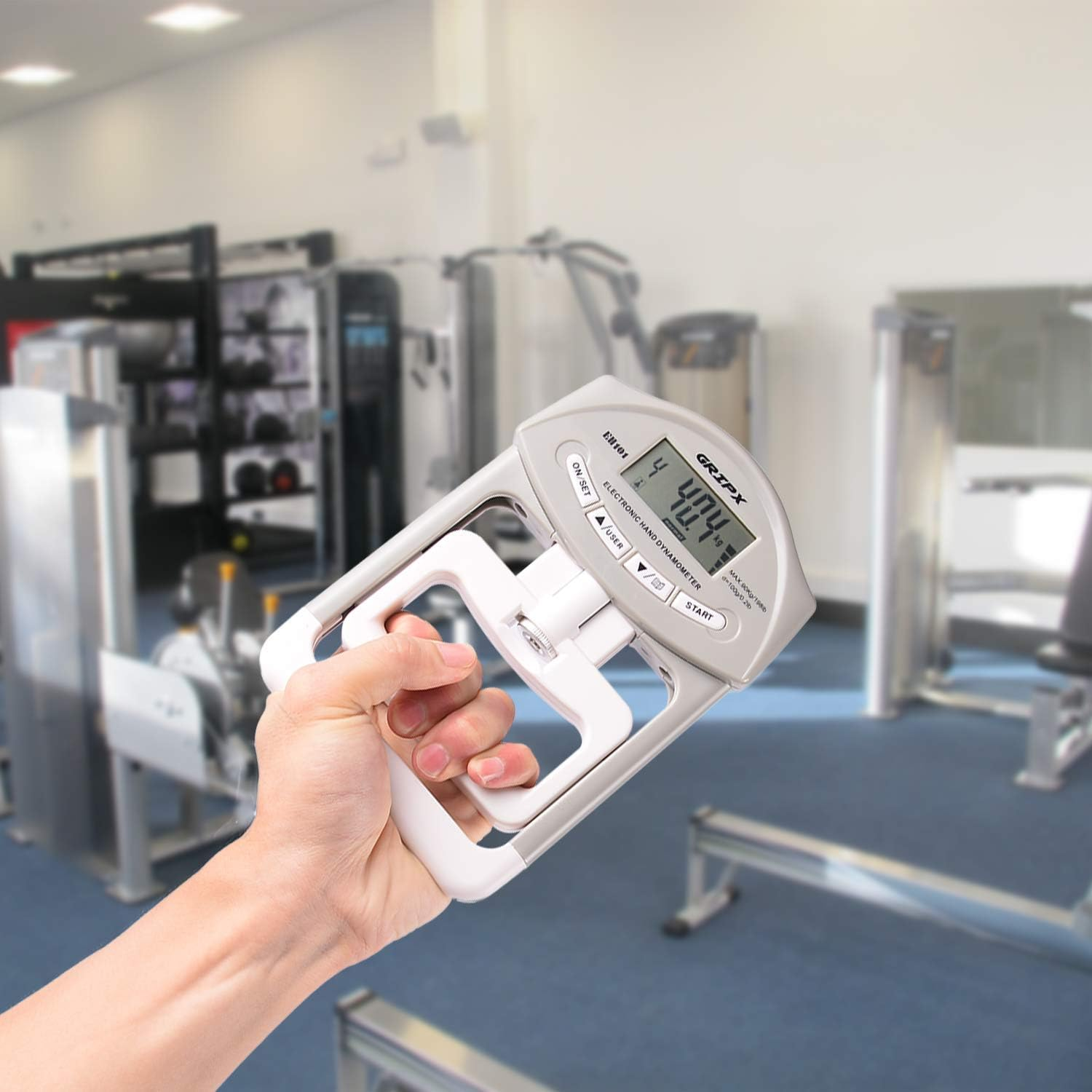 Eine Hand hält einen digitalen Handkraftmesser vor Fitnessgeräten in einem Fitnessstudio.