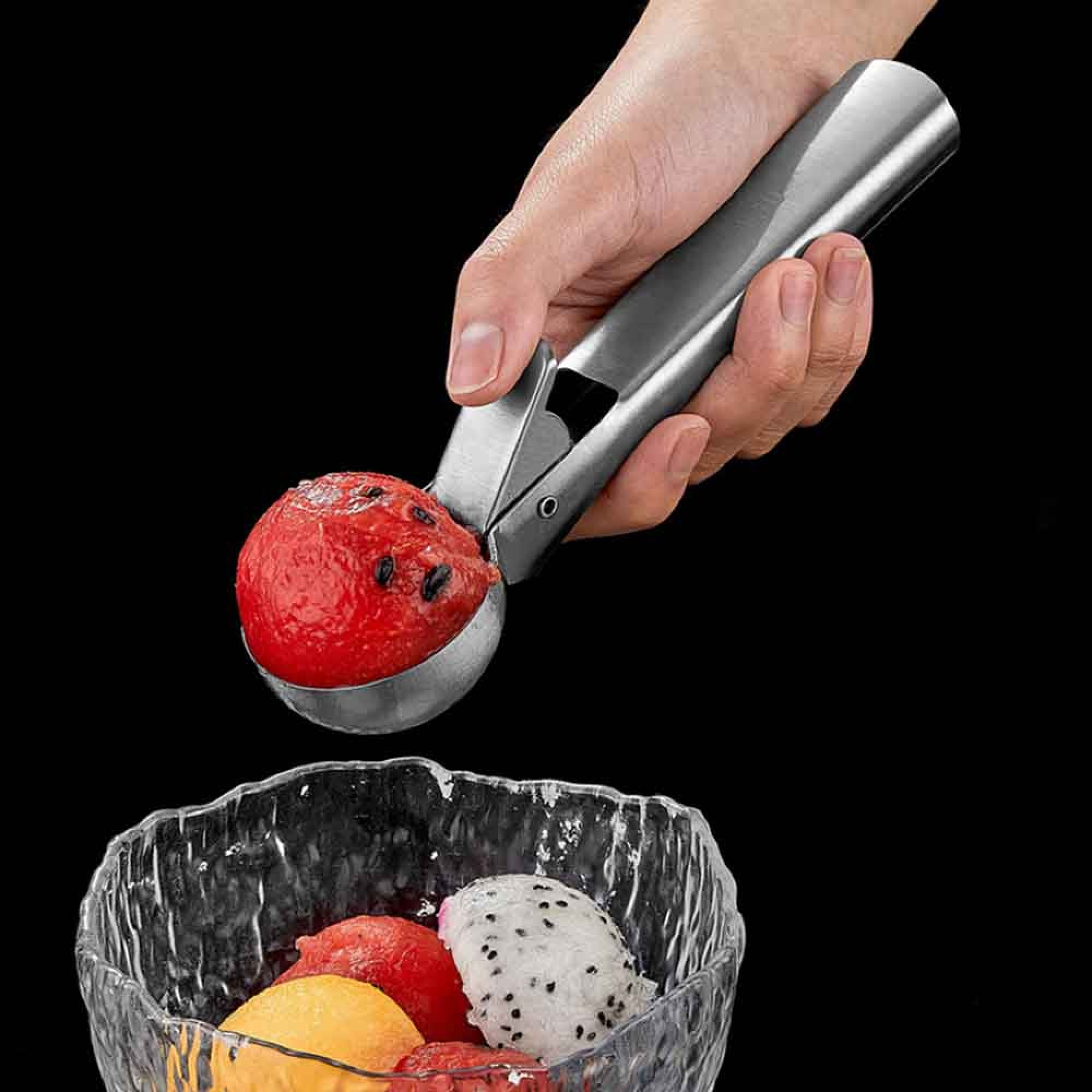 Hand schöpft rote Frucht mit schwarzen Kernen mit einem Metall-Eiskugelformer in eine Glasschale mit Früchten.