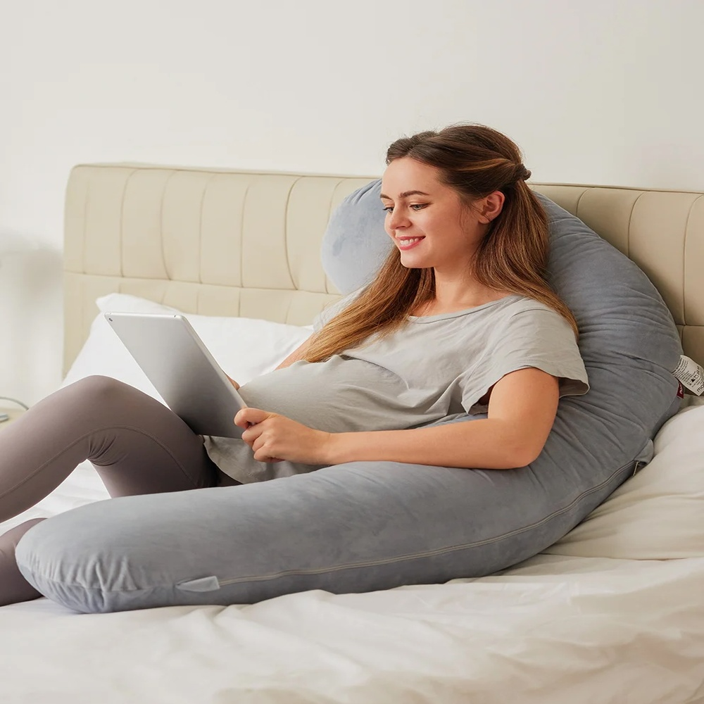 Une femme enceinte en t-shirt gris et legging utilise une tablette allongée sur un lit avec un grand coussin de corps gris.