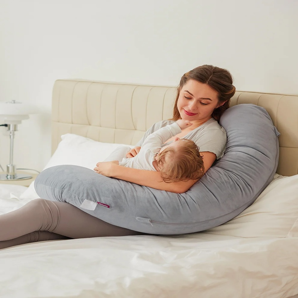 Une femme tient un bébé sur un coussin d'allaitement gris dans un lit.