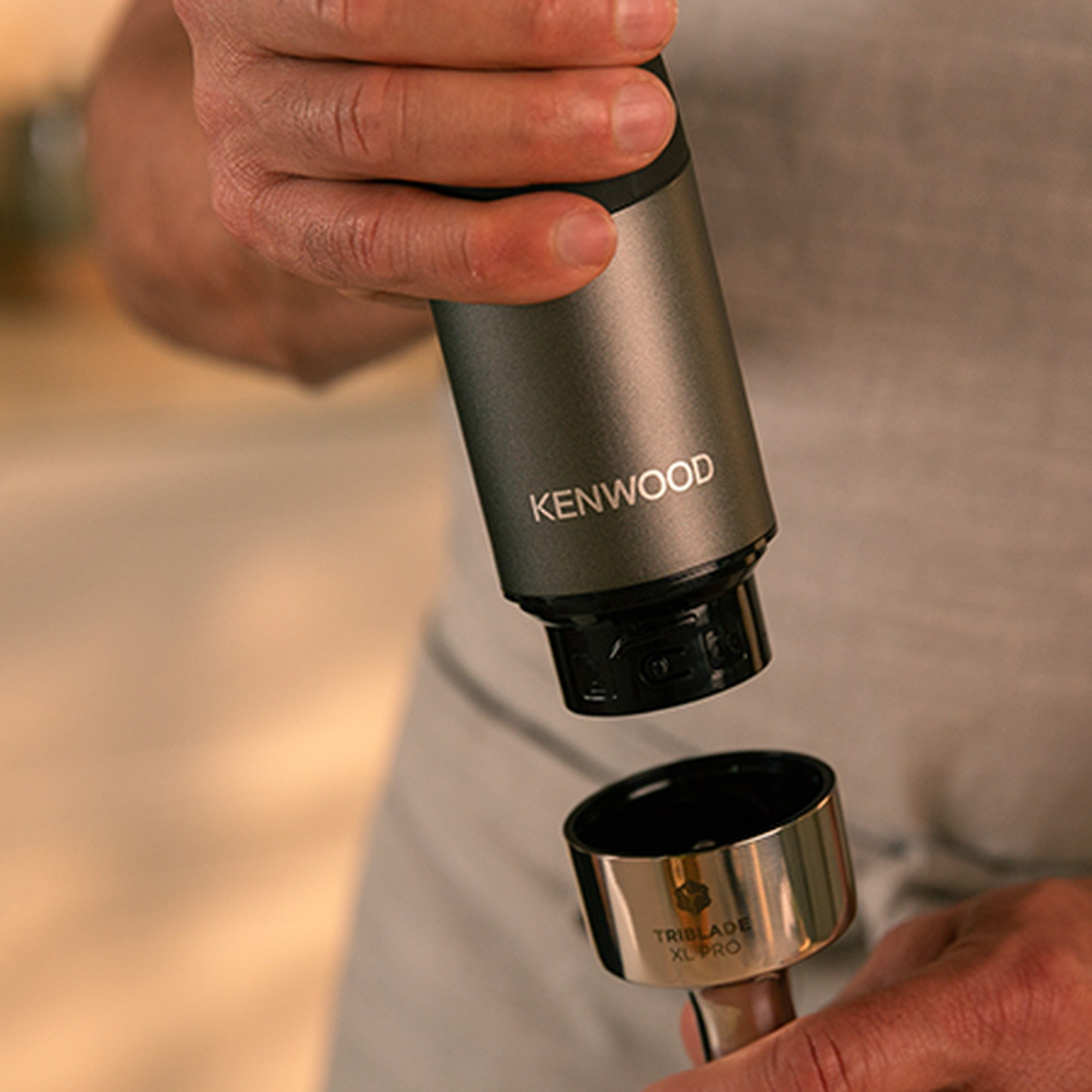 Una persona tiene un frullatore a immersione Kenwood grigio, agganciandolo all'accessorio Triblade XL Pro.