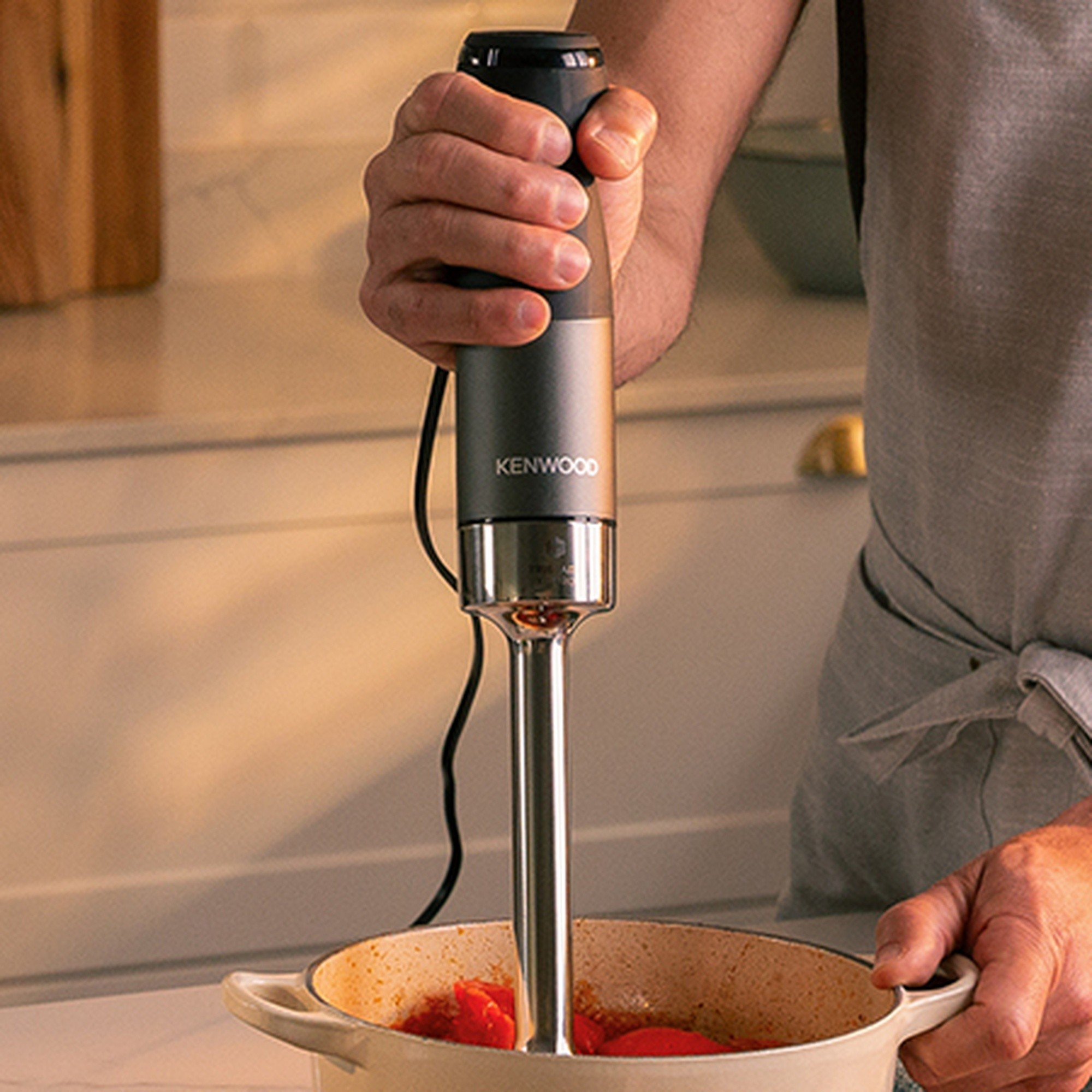 Una mano tiene un frullatore a immersione Kenwood in una pentola di pomodori.