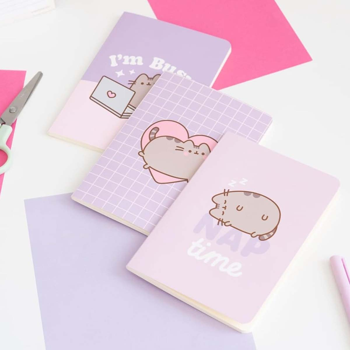 Drei Notizbücher mit Pusheen-Katzenmotiven auf lila und rosa Untergrund.