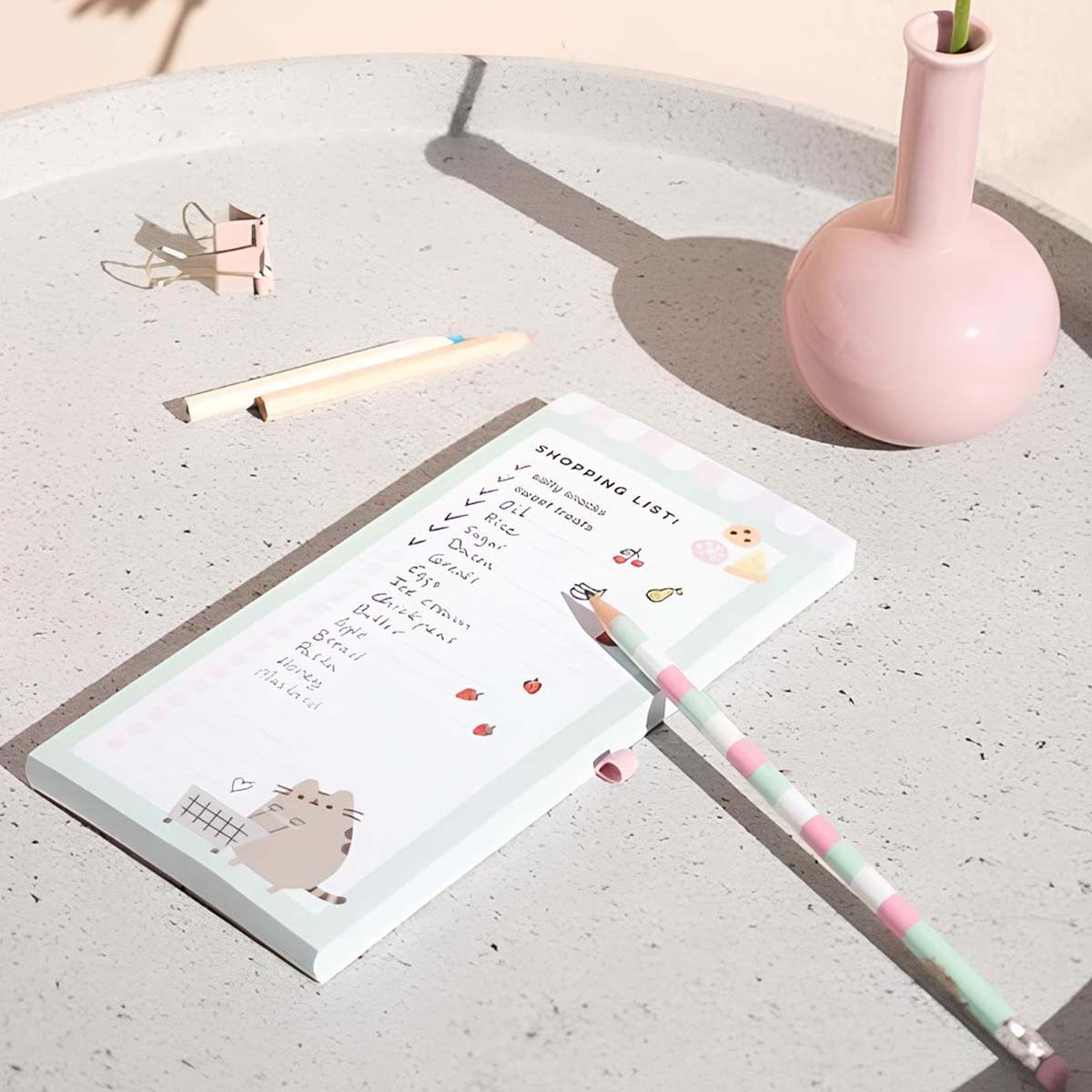 Eine Einkaufsliste mit einem Kätzchenbild, Bleistiften und einer rosa Vase.