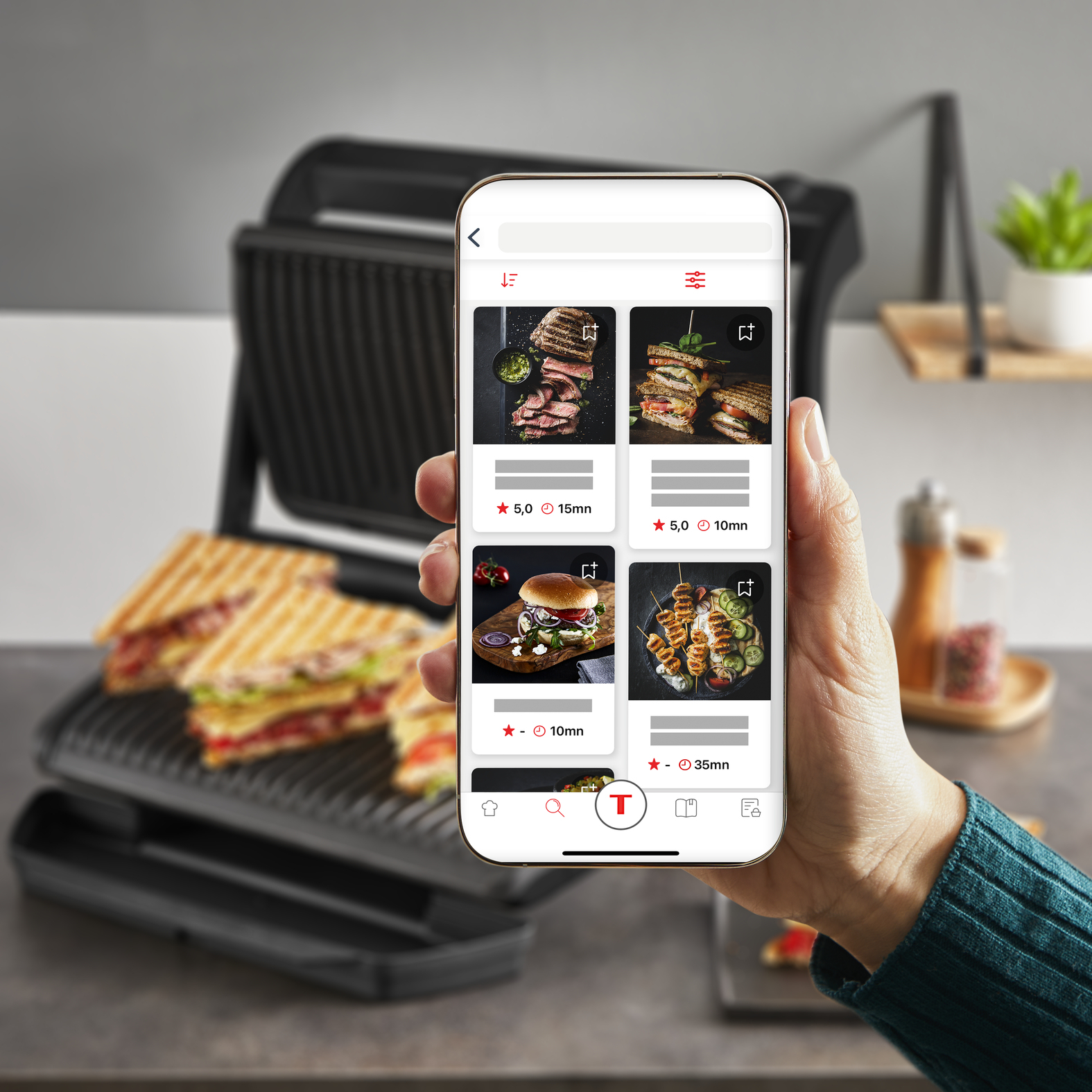 Eine Hand hält ein Smartphone mit einer Rezept-App. Ein Grill ist im Hintergrund.