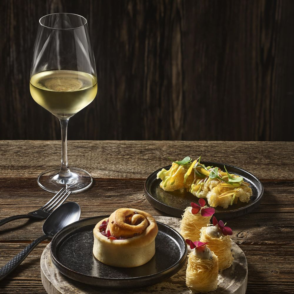 Ein Weinglas mit Weißwein, ein Brötchen und zwei kleine Desserts auf einem Holztisch.