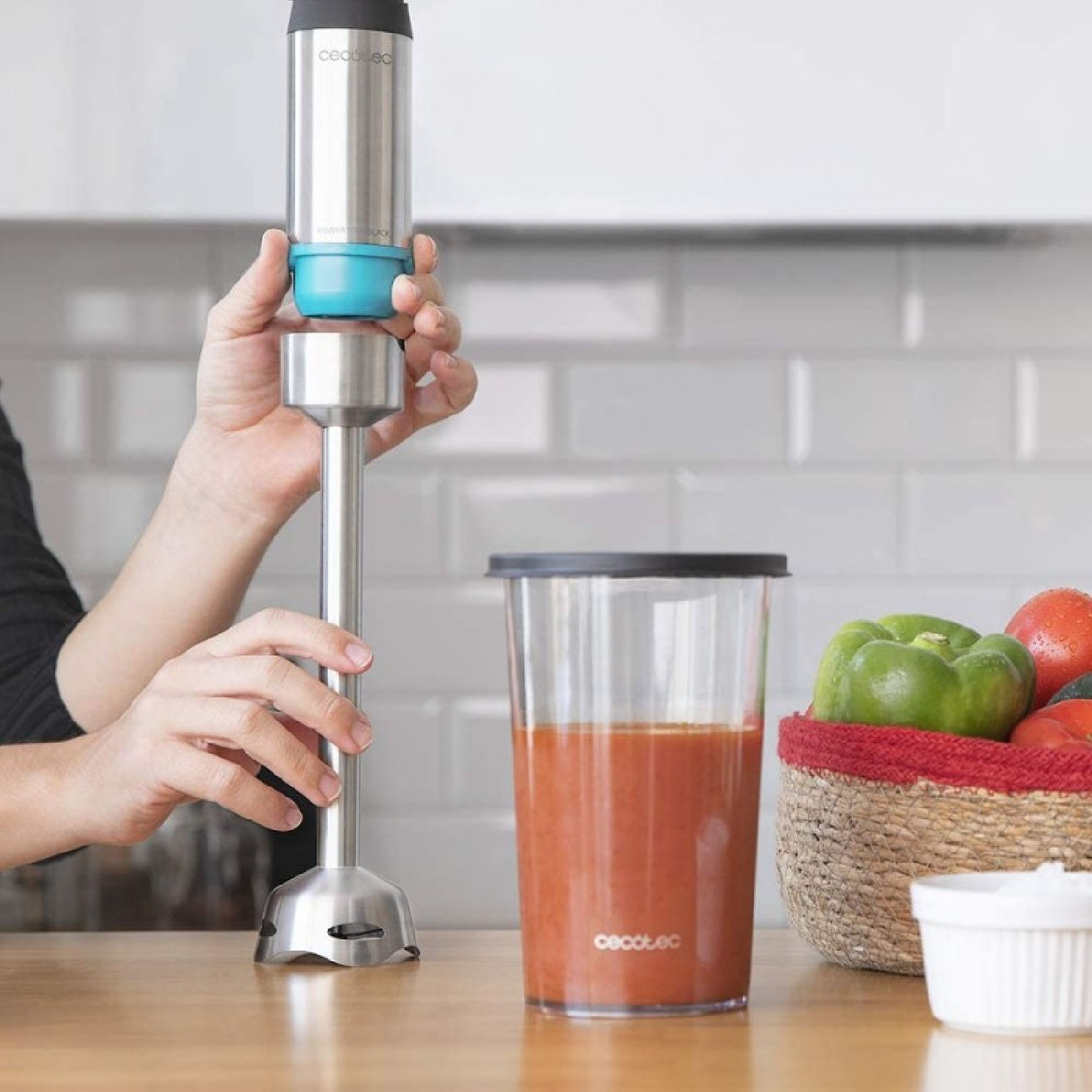 Manos ensamblan una licuadora de inmersión plateada junto a una taza de sopa de tomate y una cesta de verduras.