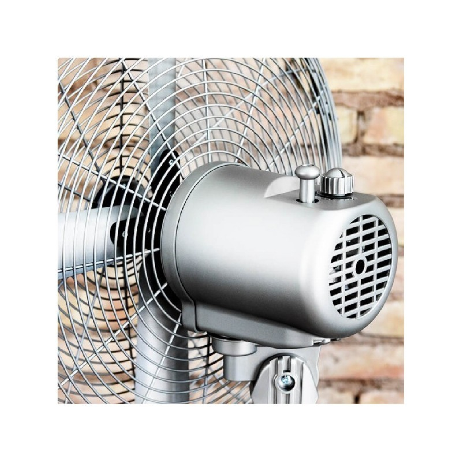 Primer plano de un ventilador eléctrico plateado con rejilla metálica y carcasa del motor contra una pared de ladrillos.