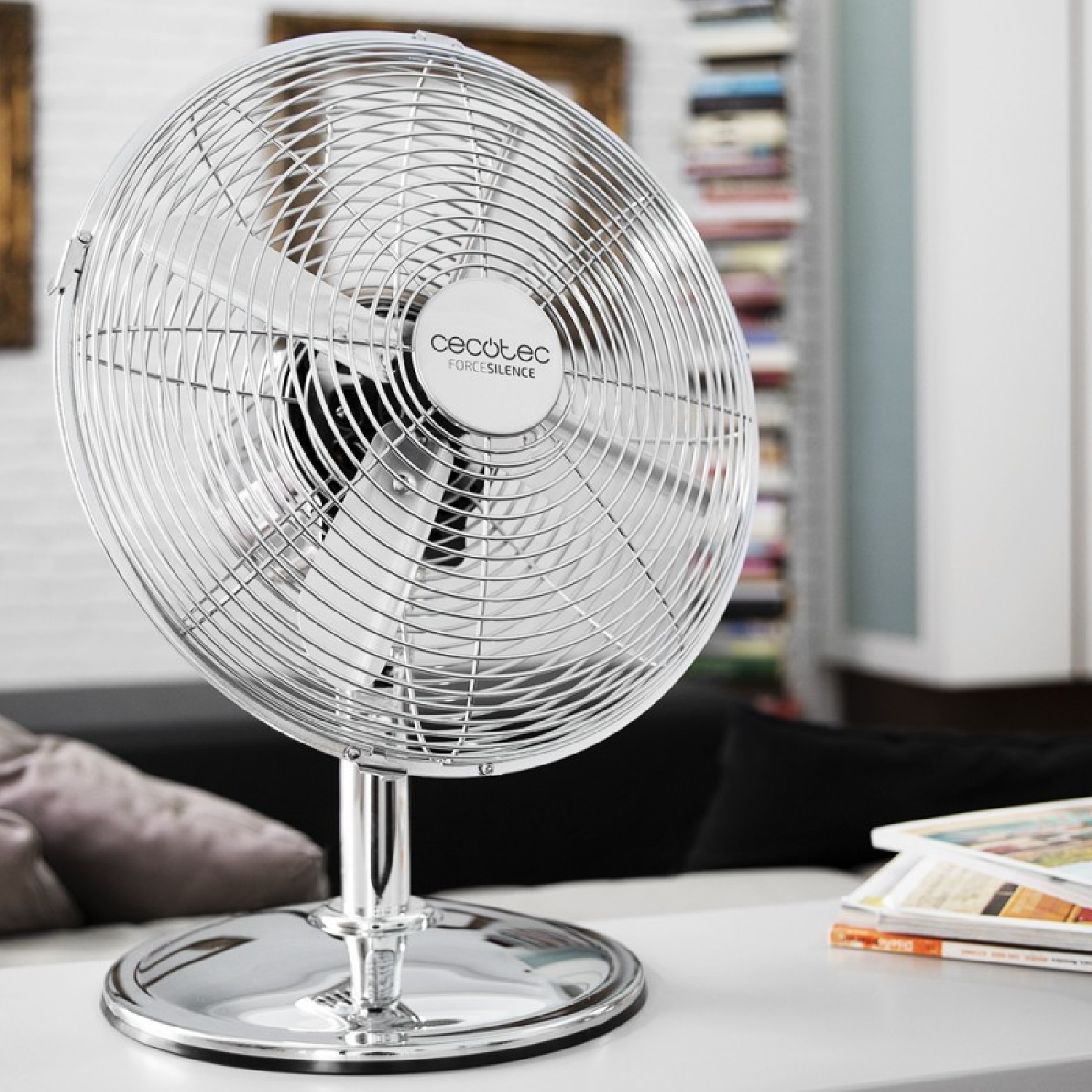 Un ventilador plateado con logo Cecotec sobre una mesa. Fondo con sofá y estantería.