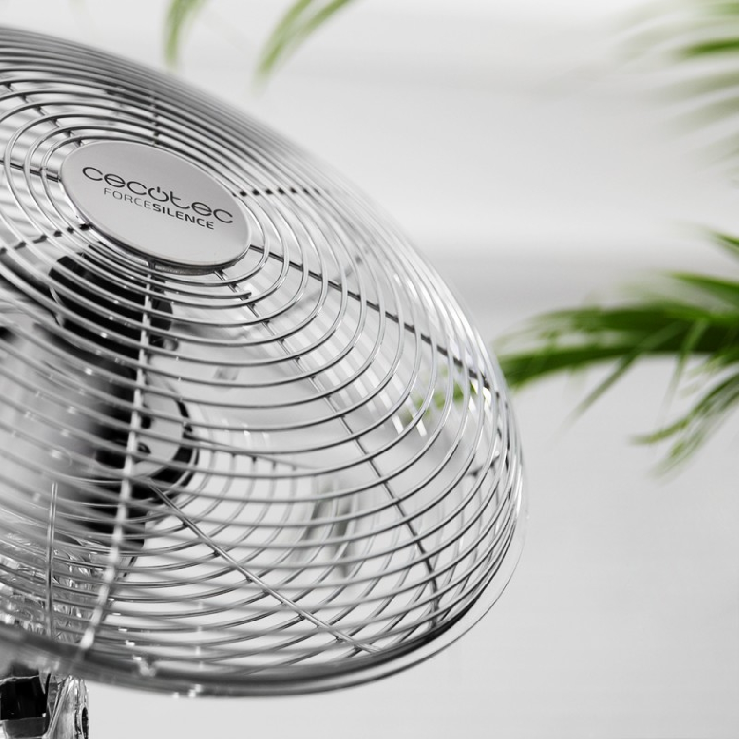 Primer plano de una rejilla de ventilador plateada con una planta al fondo borroso.