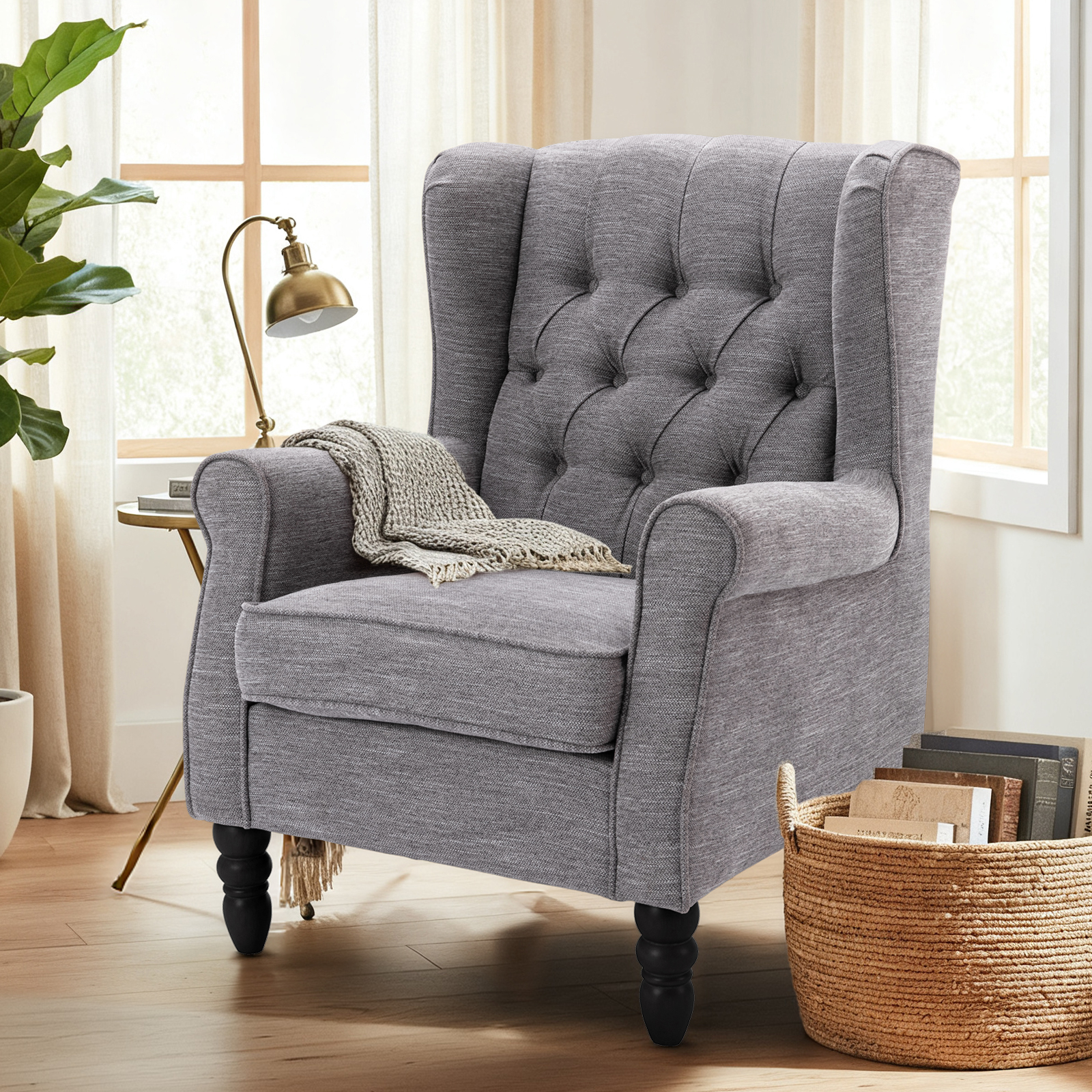 Poltrona grigia con bottoni, coperta, lampada, cesto con libri e pianta.