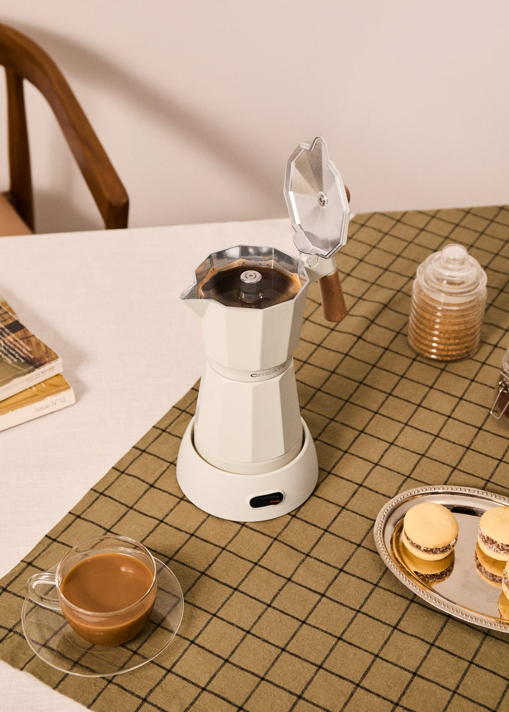 Una cafetera blanca con la tapa abierta, junto a una taza de café y galletas.
