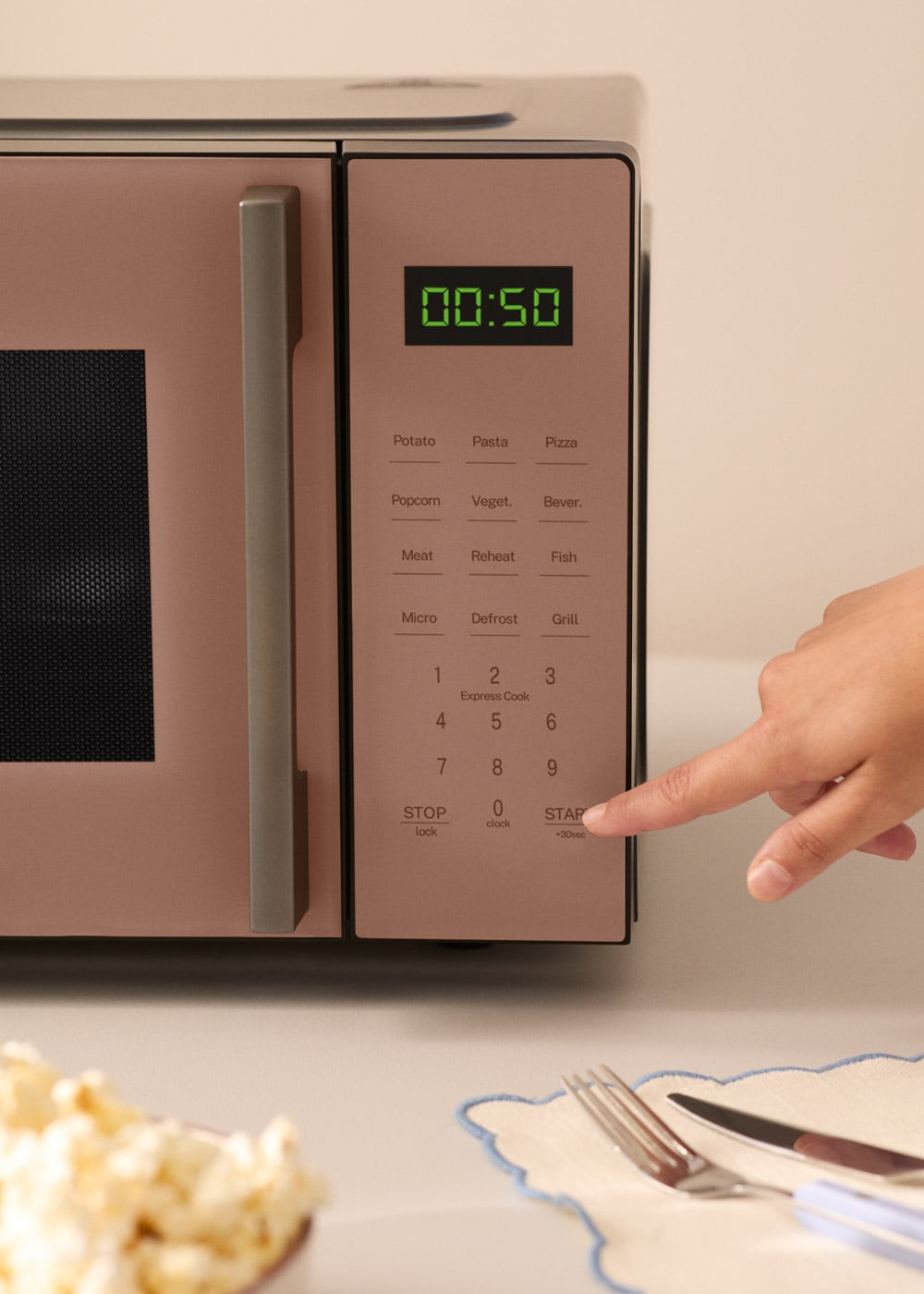 Una mano presiona el botón de inicio en un microondas oro rosa con pantalla digital que muestra 00:50.