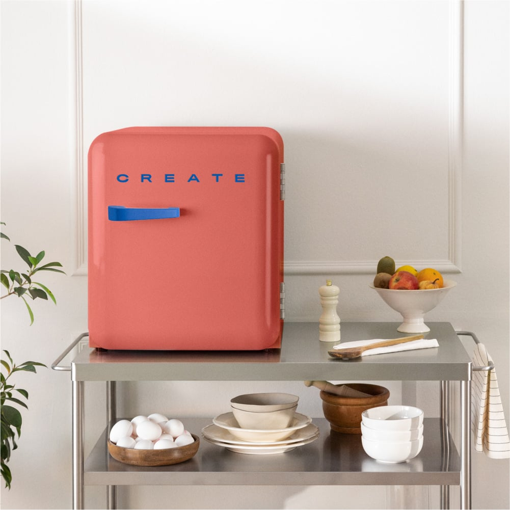 Una mini nevera coral con 'CREATE' escrito encima está en un carrito metálico con frutas y huevos.