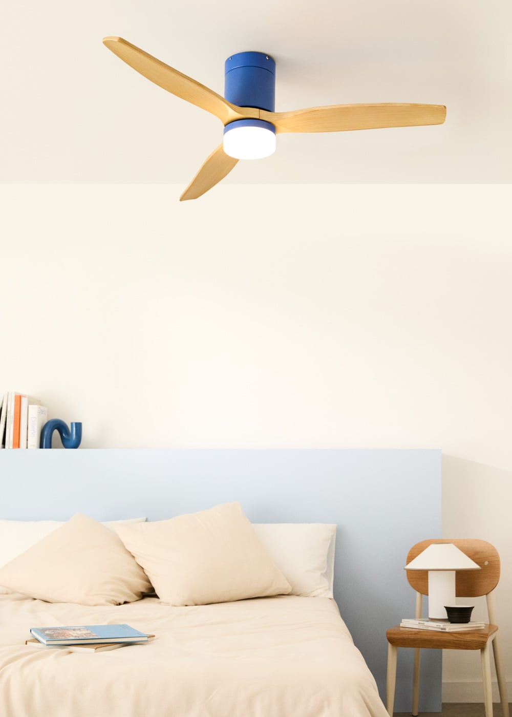 Un ventilador de techo azul con aspas de madera sobre una cama con almohadas beige.