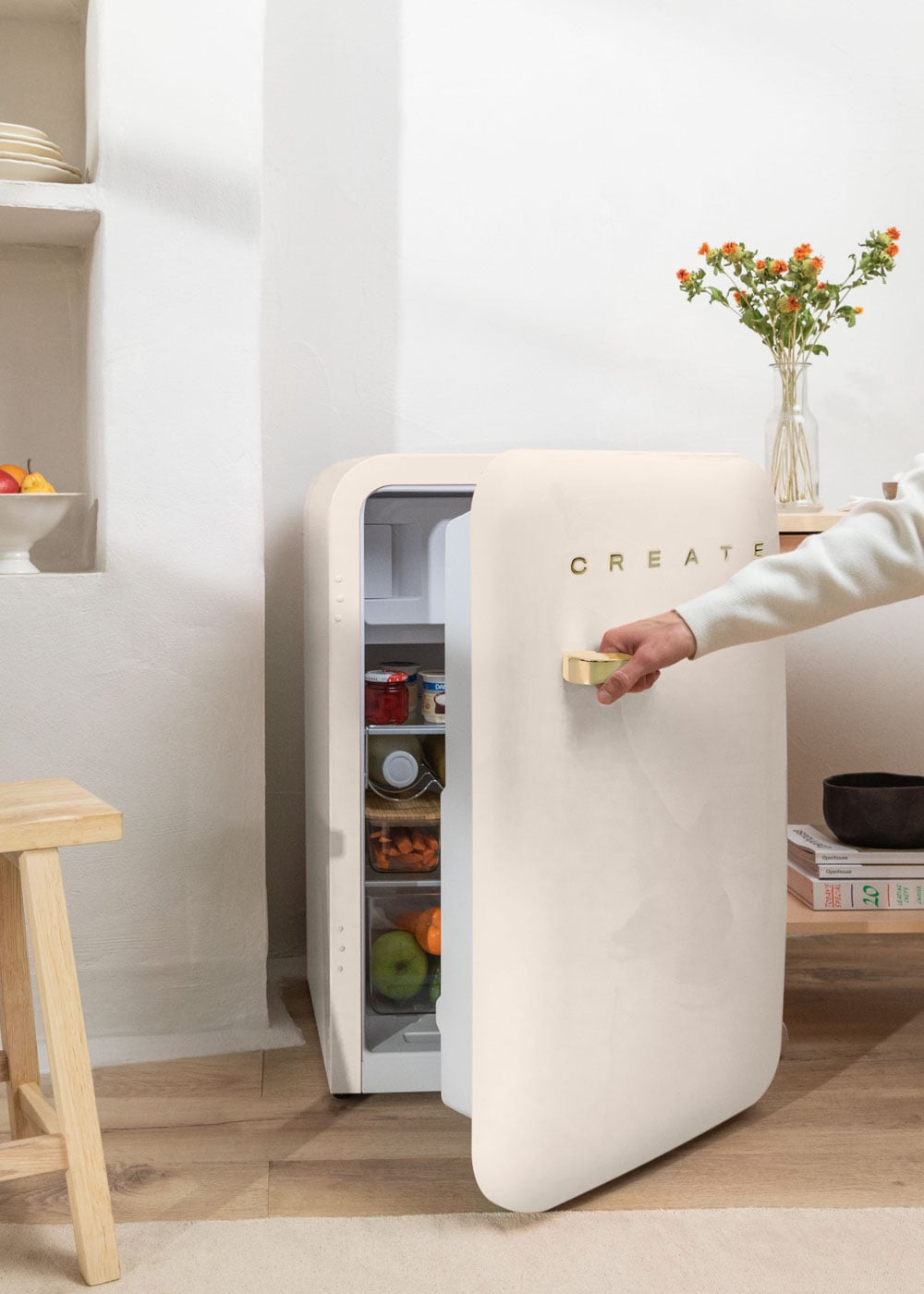 Una mano abre un refrigerador color crema con un tirador dorado. Se ven alimentos en el interior.