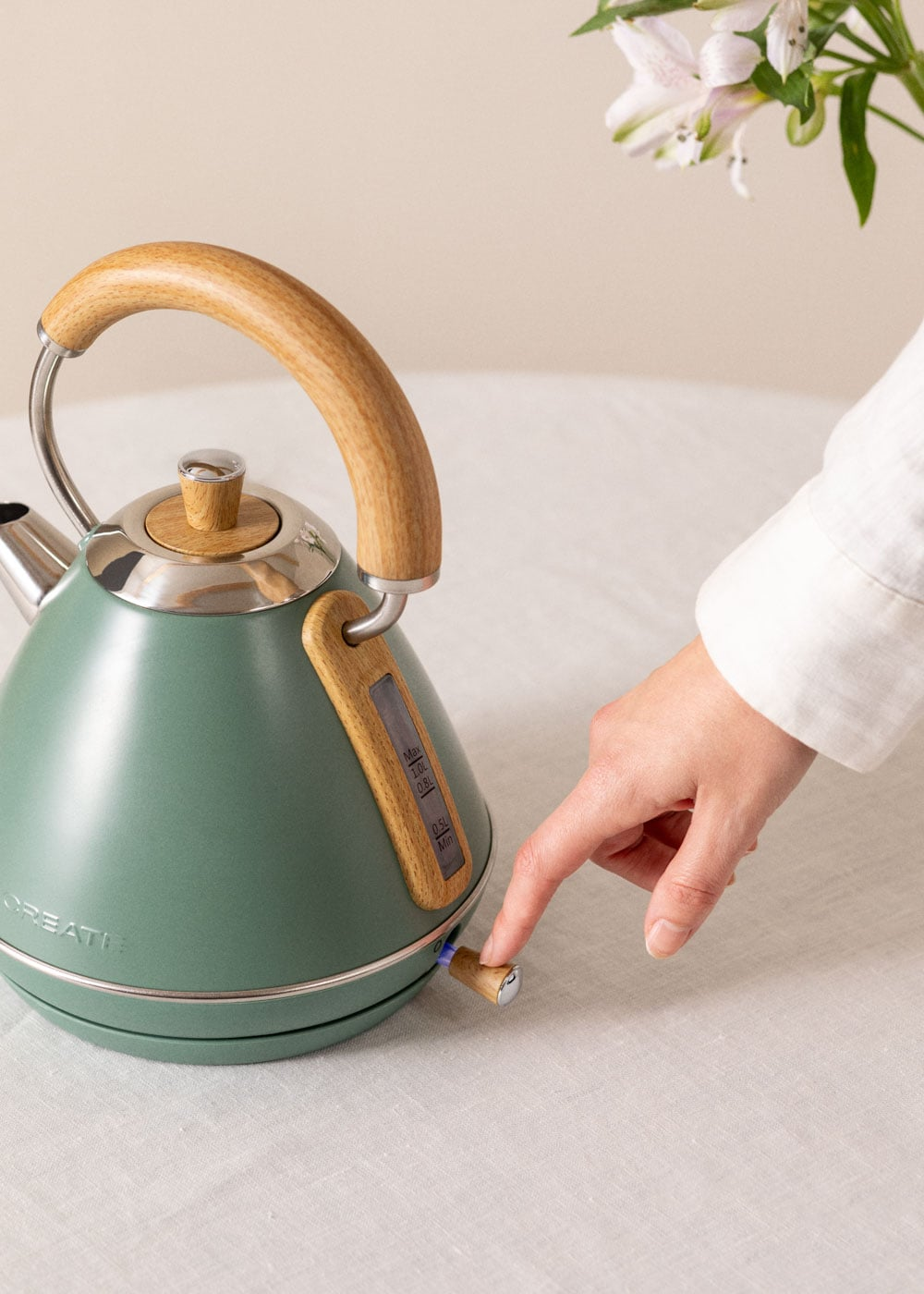 Un hervidor verde con mango y detalles de madera. Una mano presiona el interruptor.
