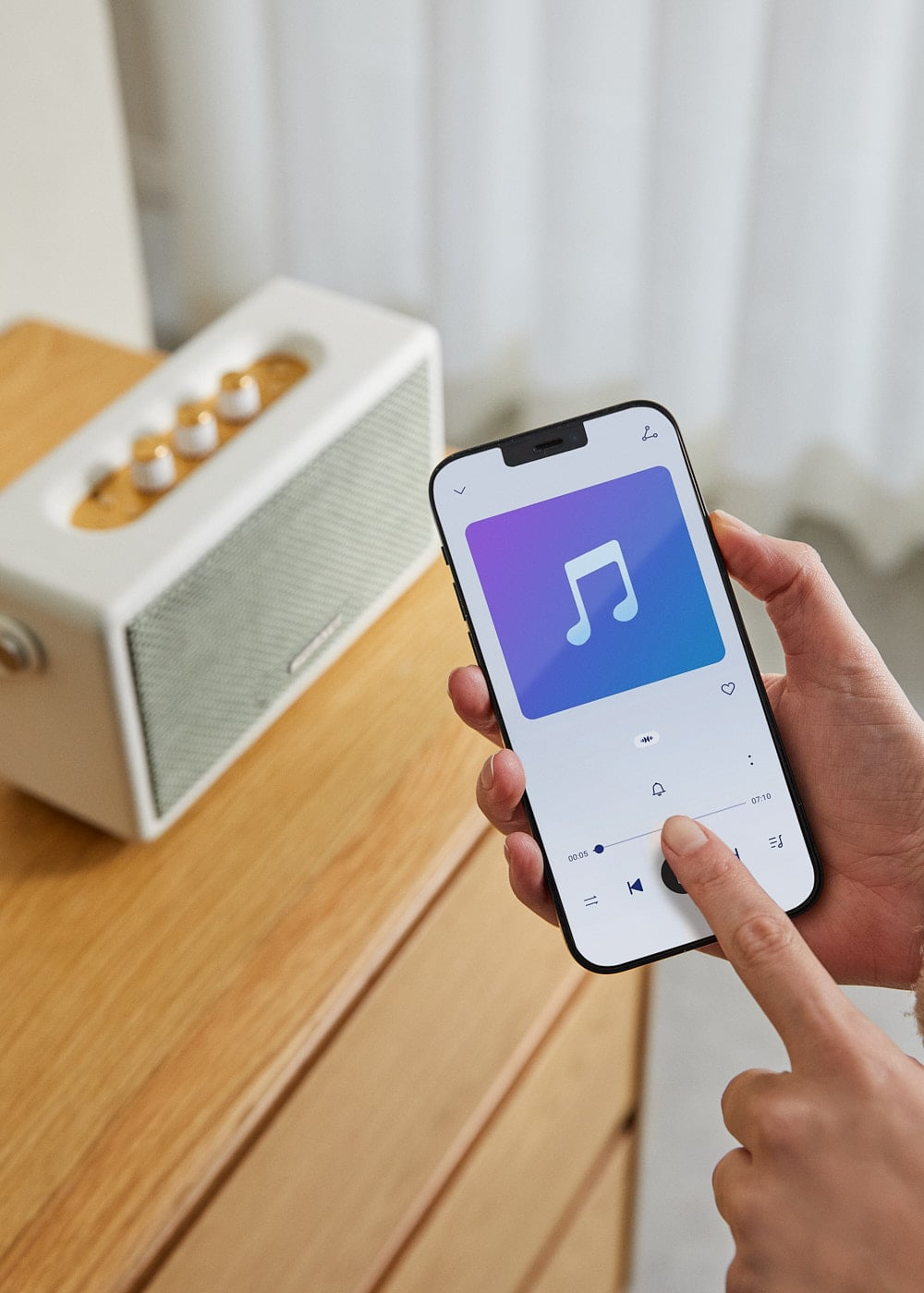 Una mano sostiene un smartphone con app de música, al lado un altavoz blanco.