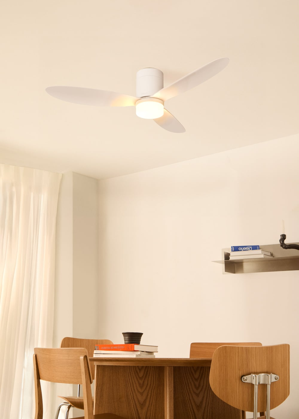 Un ventilador de techo blanco con tres aspas y una lámpara en el centro, iluminando una habitación con una mesa y sillas de madera.