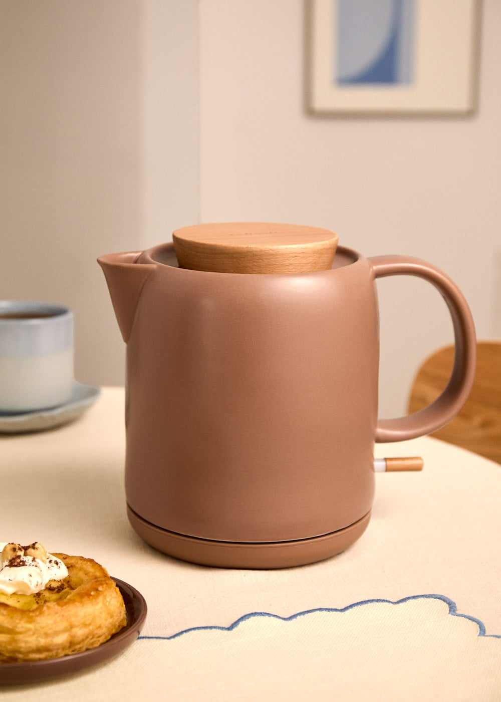 Una tetera marrón con tapa de madera está sobre una mesa con pasteles y una taza.