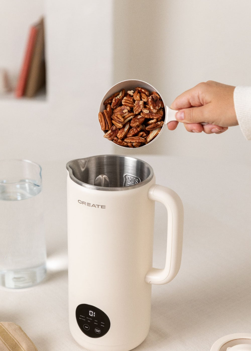 Una mano vierte nueces pecanas en una lechera blanca junto a un vaso de agua.