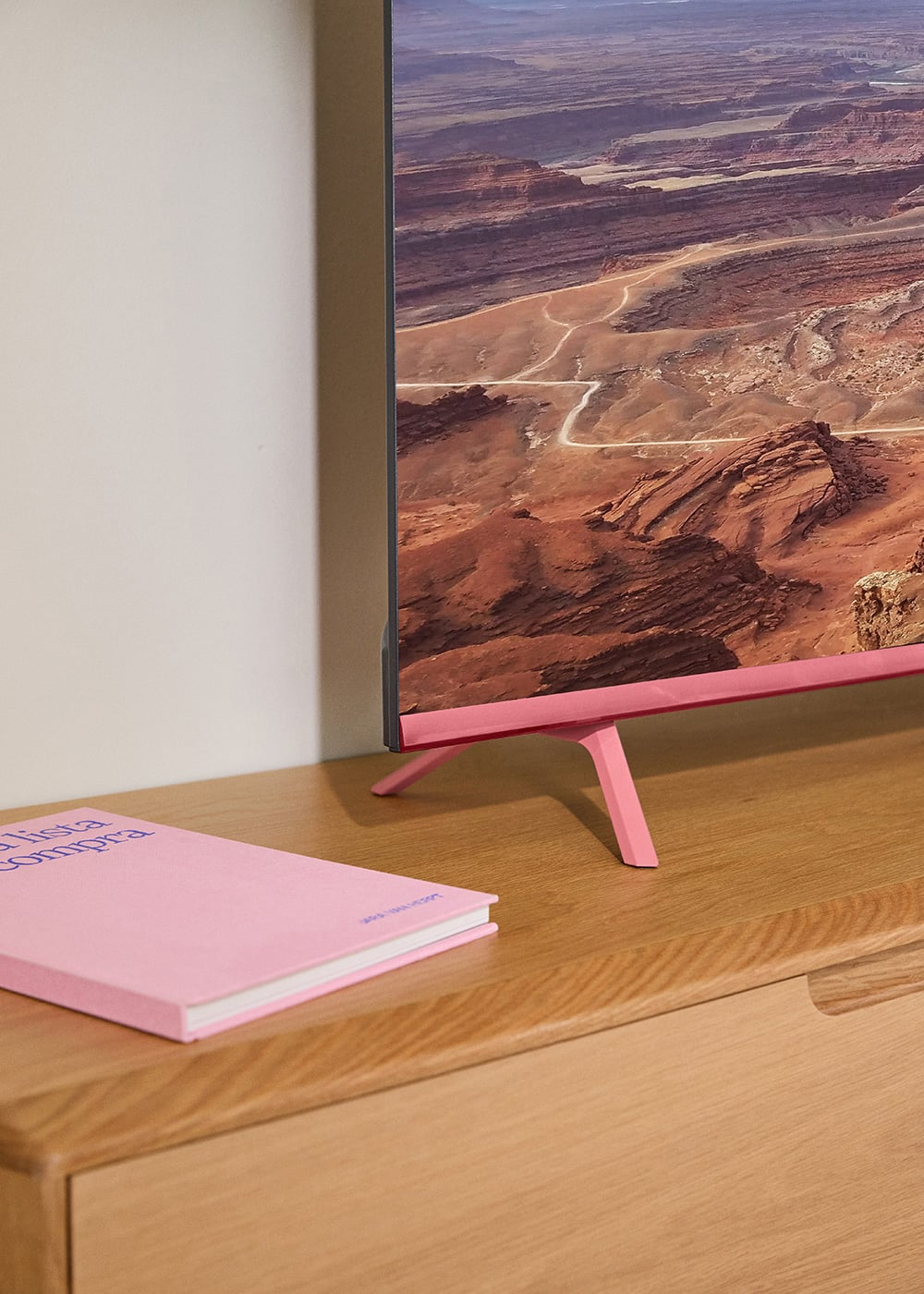 Televisor rosa con un paisaje en pantalla, junto a un libro rosa sobre mesa de madera.