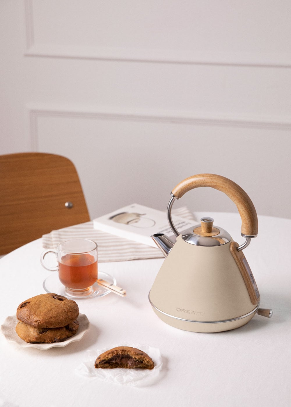 Una tetera beige con mango de madera sobre mesa blanca con galletas y té.
