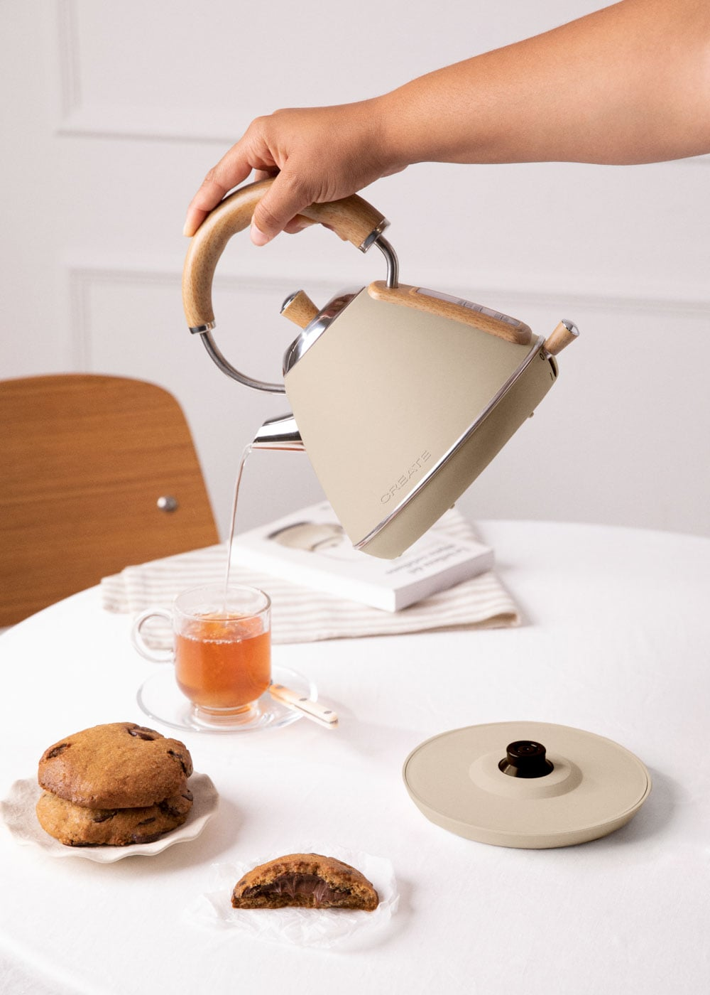Una mano vierte té marrón de una tetera color crema en una taza de vidrio sobre una mesa blanca.