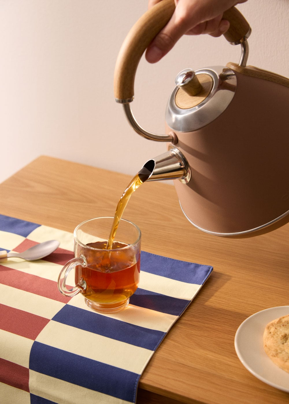Tetera marrón vertiendo té en una taza de vidrio sobre un camino a cuadros.