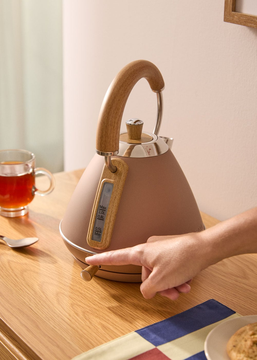 Tetera marrón con asa de madera, dedo accionando interruptor. Taza de té al lado.