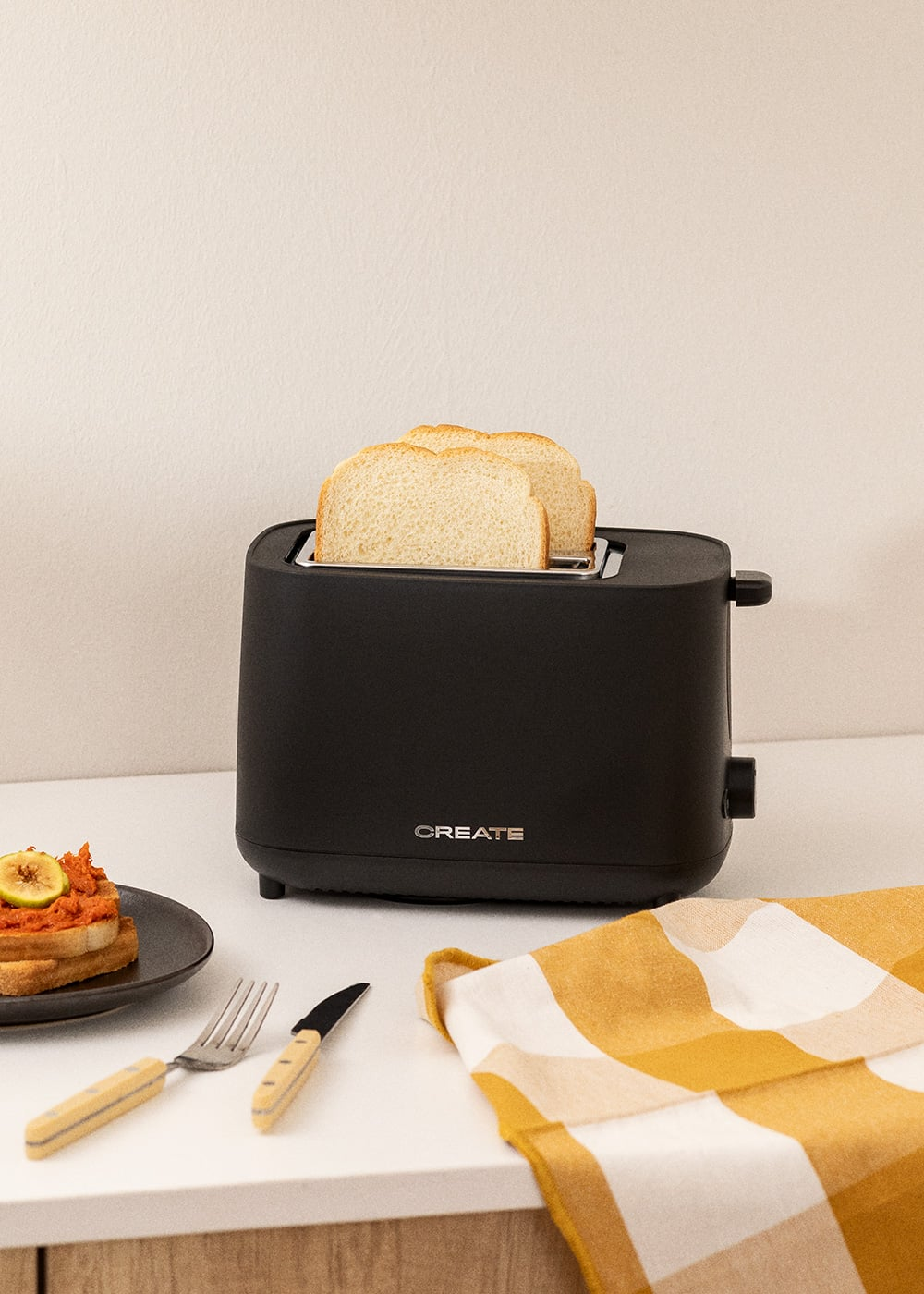 Un grille-pain noir avec deux tranches de pain, une assiette de toast et un torchon à carreaux jaune et blanc.