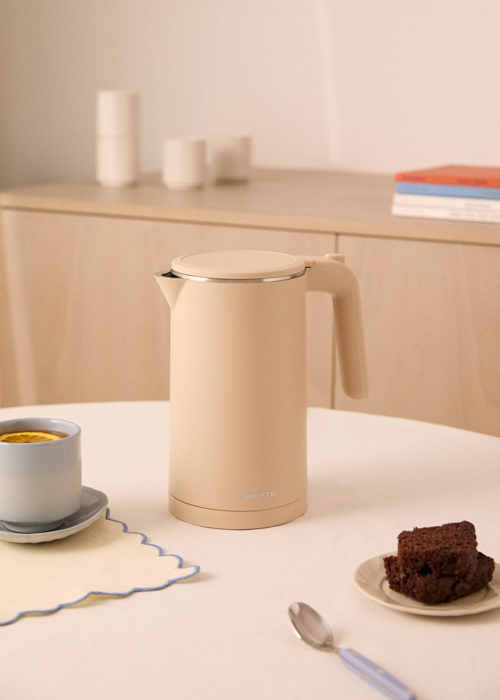 Un hervidor beige está sobre una mesa con una taza de té y brownies.