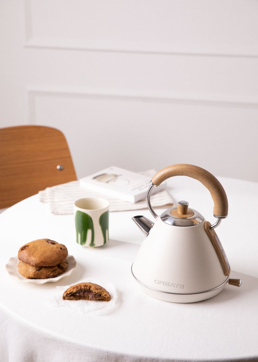 Una tetera crema con mango de madera, galletas y una taza sobre una mesa.