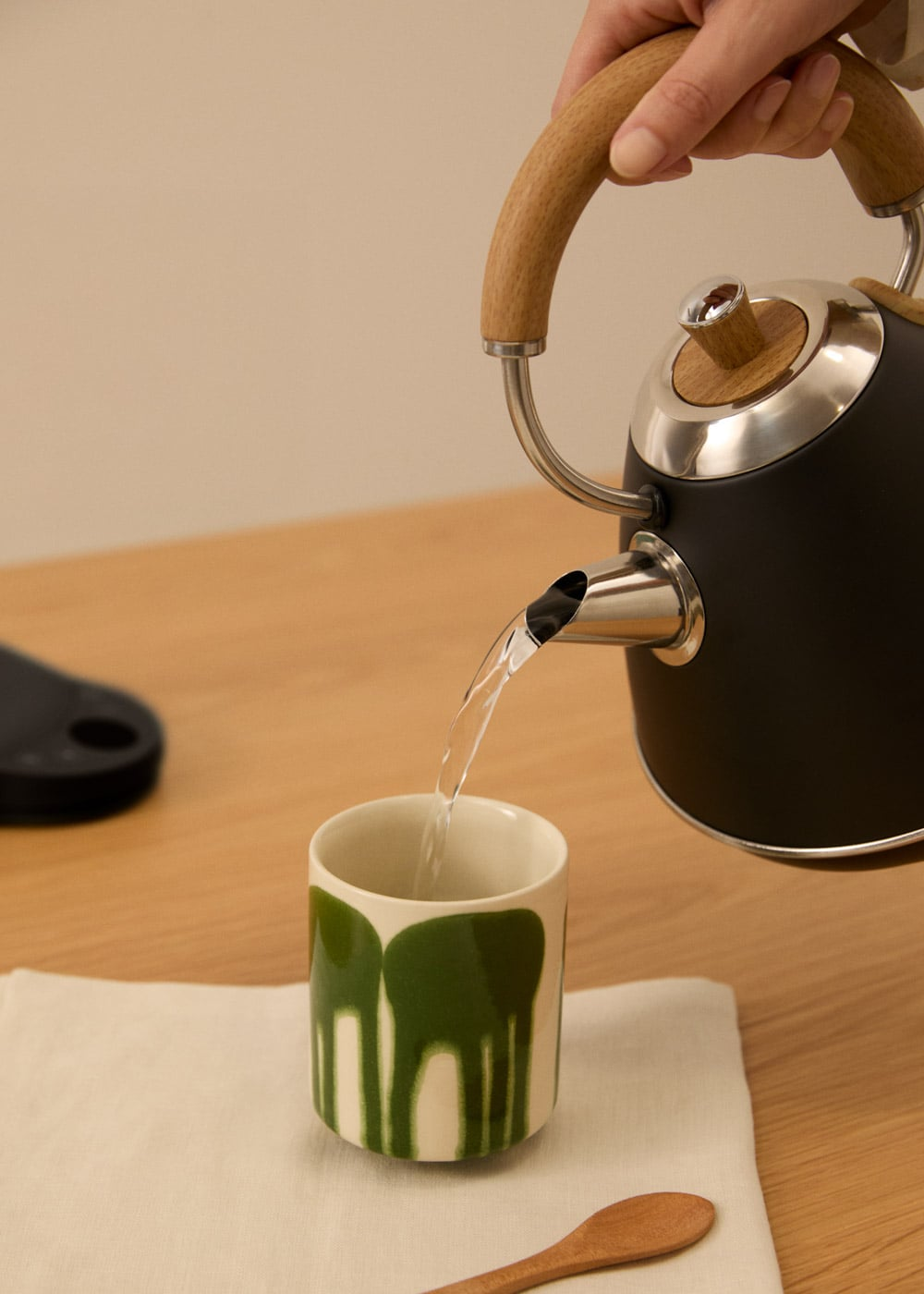 Una mano vierte agua de una tetera negra con mango de madera en una taza verde.