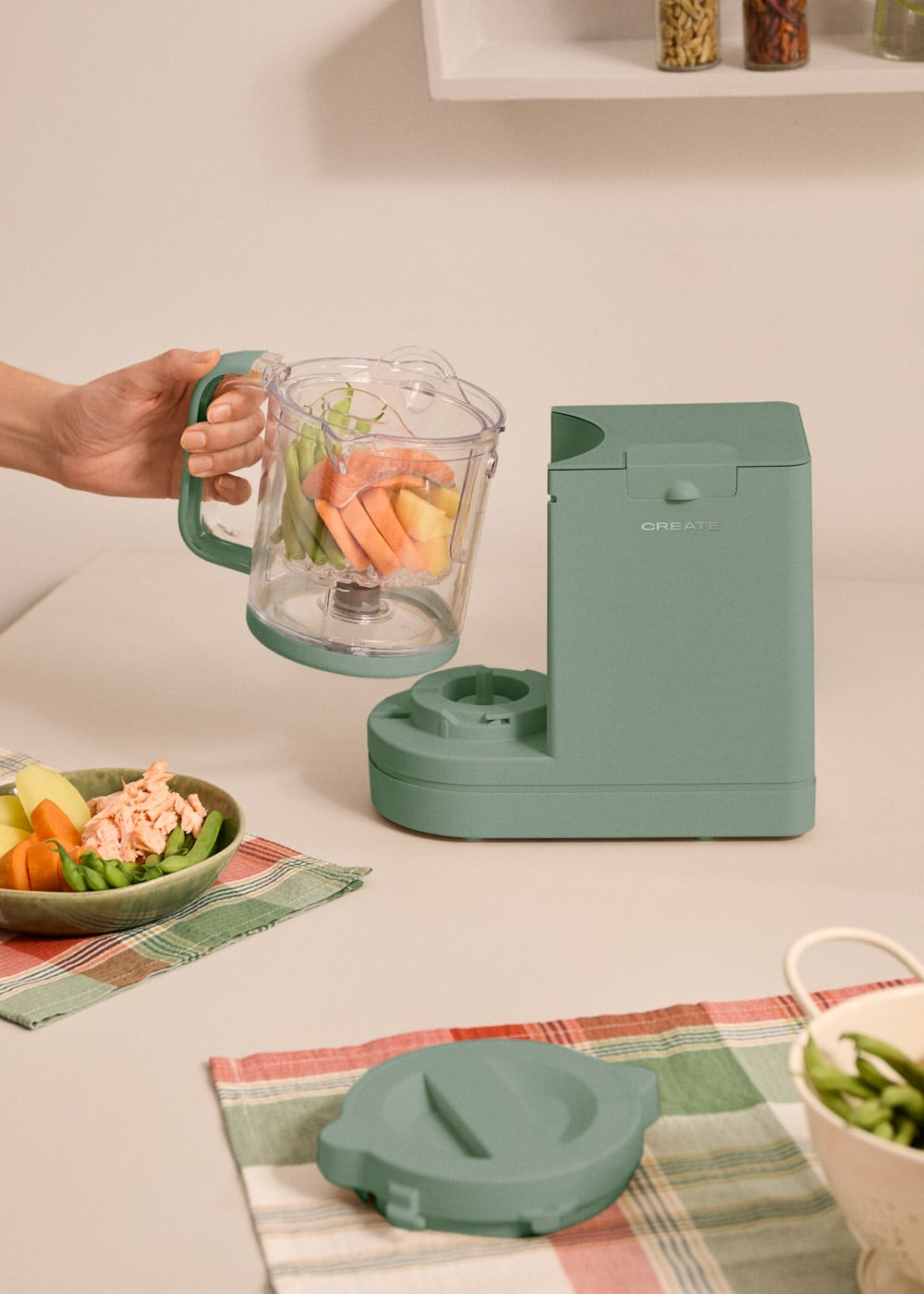 Una mano sostiene un vaso de batidora con verduras; un cuenco con comida está cerca.