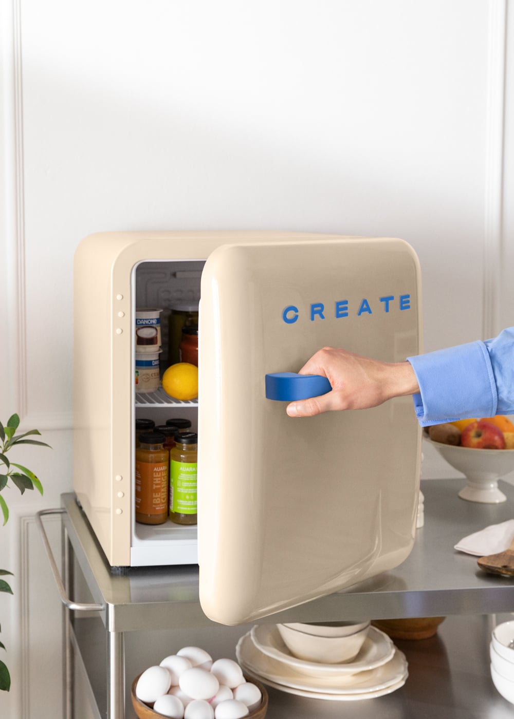 Una mini nevera color crema con texto azul 'CREATE'. Una mano abre la puerta revelando el contenido.