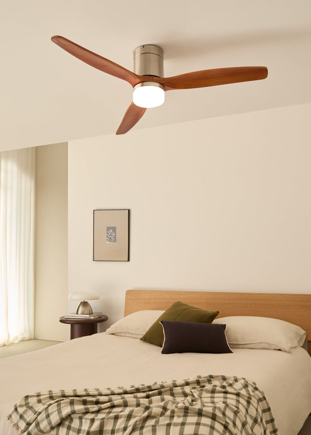 Un ventilador de techo con aspas de madera sobre una cama con cojines y manta a cuadros.