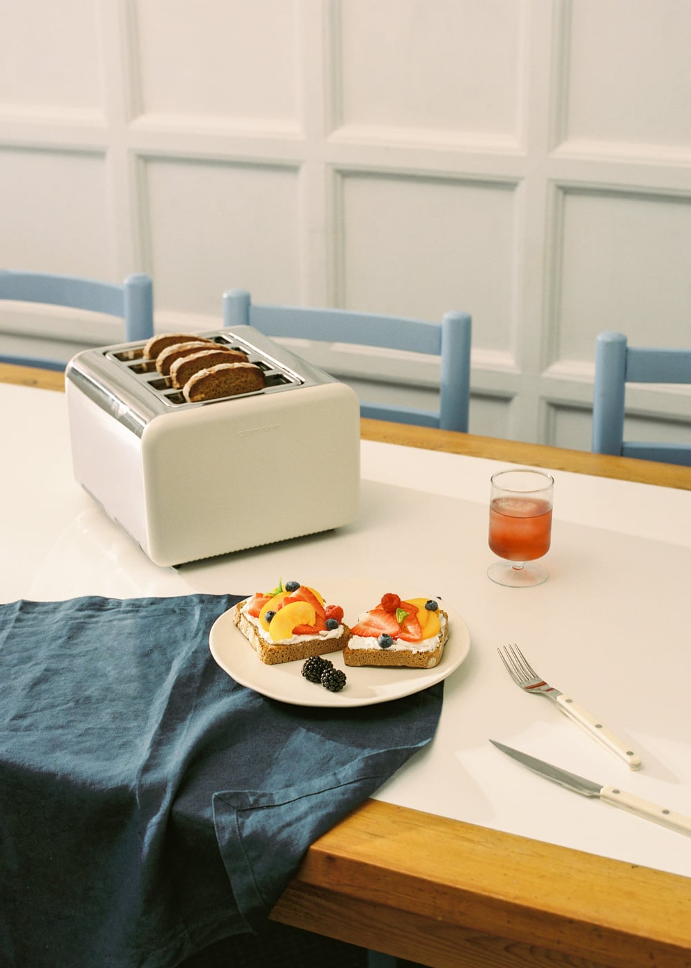 Una tostadora con pan, dos sándwiches abiertos con fruta, un vaso de zumo y cubiertos en una mesa.