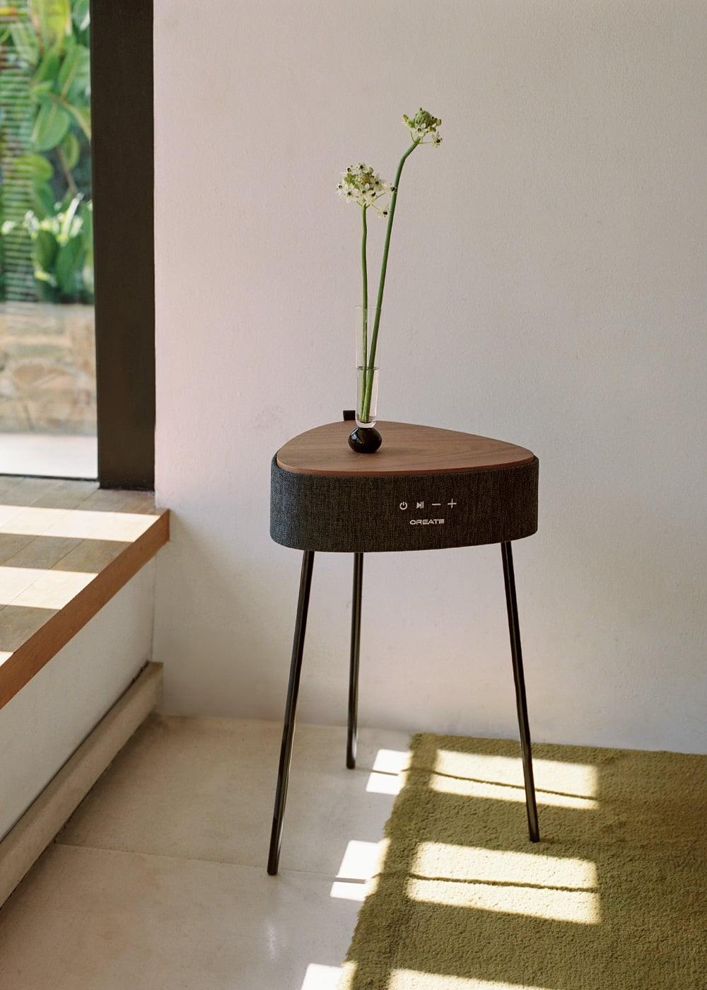 Una mesa triangular de madera con base de altavoz sostiene un jarrón de cristal con flores blancas.