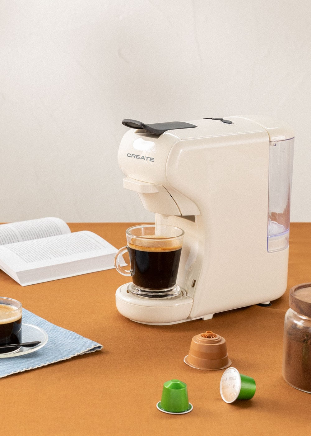 Una cafetera color crema prepara espresso en una taza de vidrio. Cápsulas de café y un libro cerca.