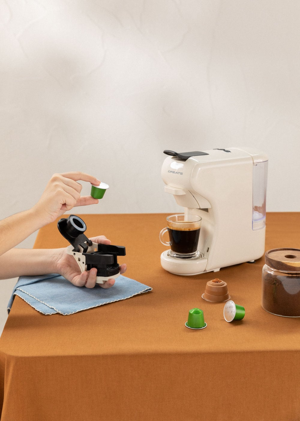 Una mano sostiene una cápsula de café verde sobre una cafetera con una taza de café.